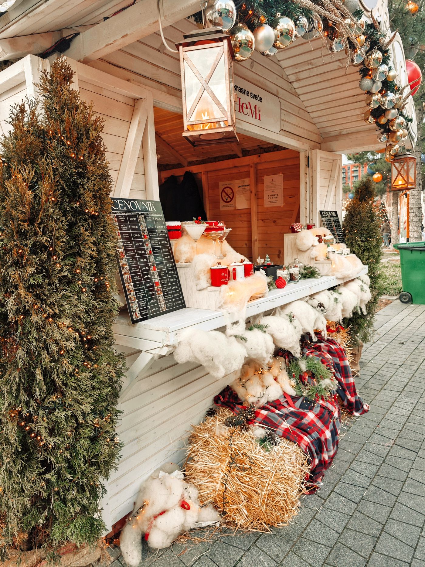 A wooden market stall displays holiday decorations, mugs and plush ornaments beside evergreen trees.