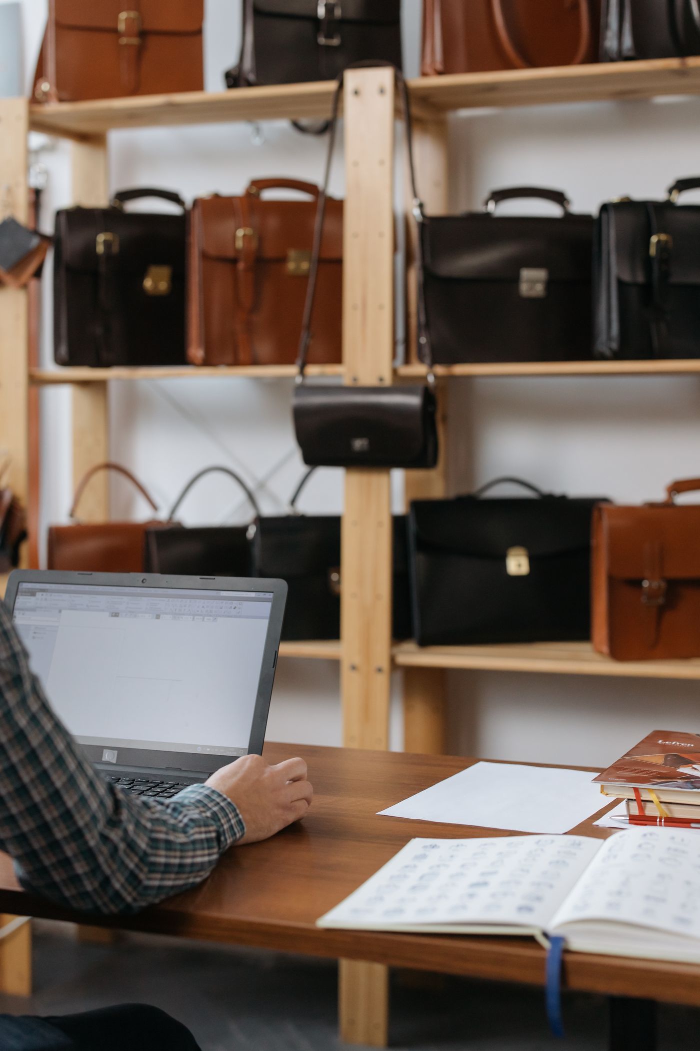 A person works at a laptop and desk in a shop with leather bags on shelves.