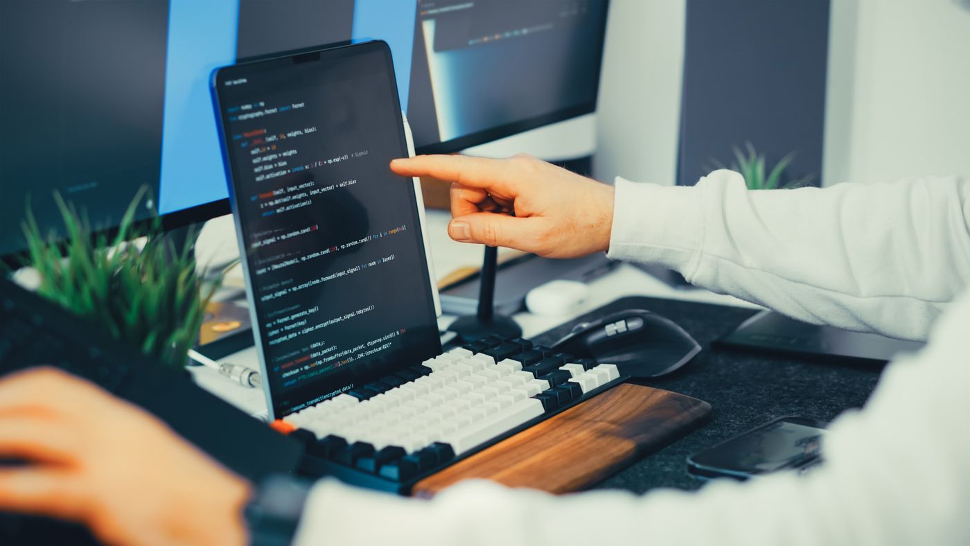 A programmer points toward code displayed on a laptop and monitor.