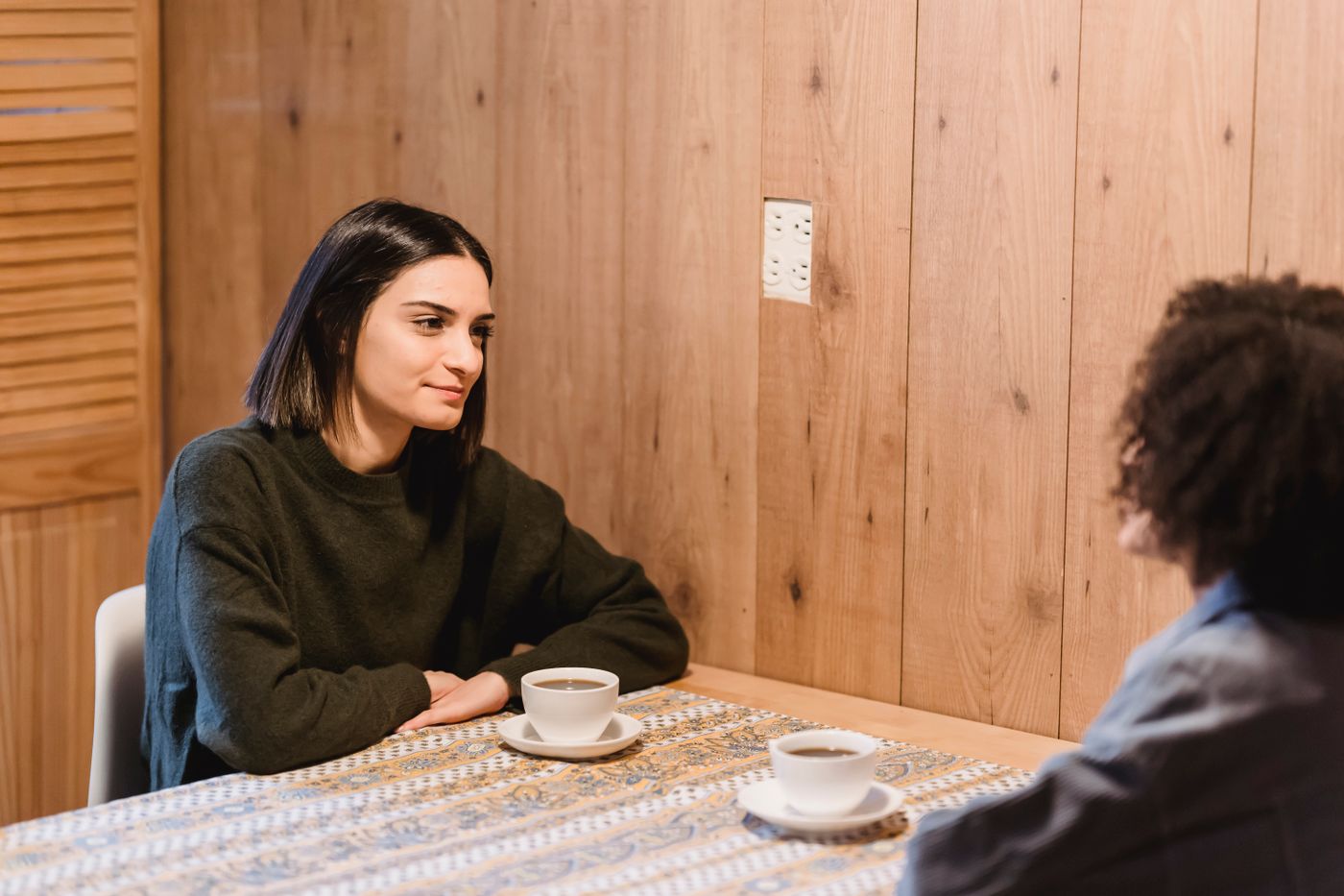 Two people talk across a café table with cups of coffee.